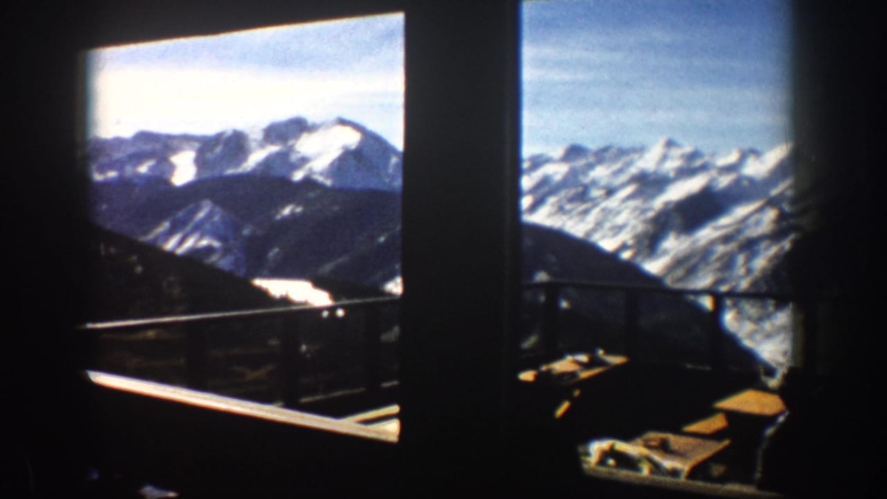 1961 Aspen home-movie frame from inside a lodge or hotel, with large windows framing snowy peaks under a blue sky.