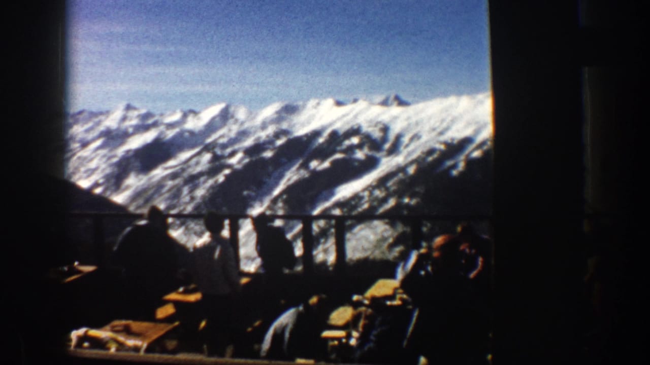 1961 Aspen home-movie frame showing dark restaurant silhouettes against bright snow-capped Colorado mountain ridges.