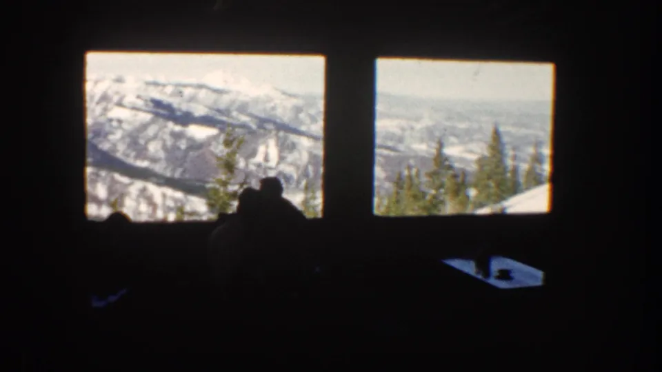 1961 Aspen lodge or restaurant view with silhouetted guests below bright windows showing snow-covered mountains and trees.