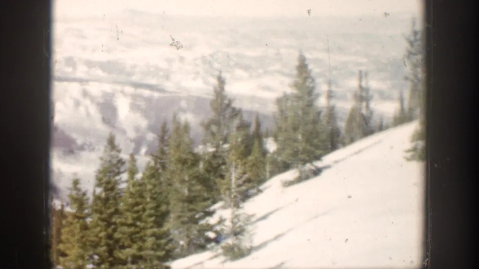 Snow-covered Aspen hillside with pine trees in the foreground and hazy mountain ridges beyond.