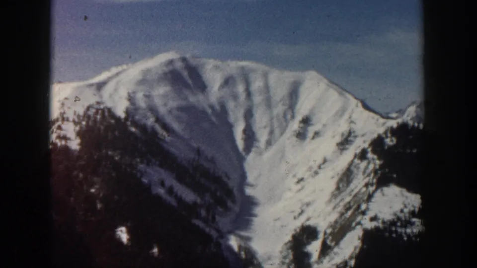 Aspen-area 1961 home-movie frame showing a snowy mountain ridge, blue sky, and dark evergreen-covered lower slope.