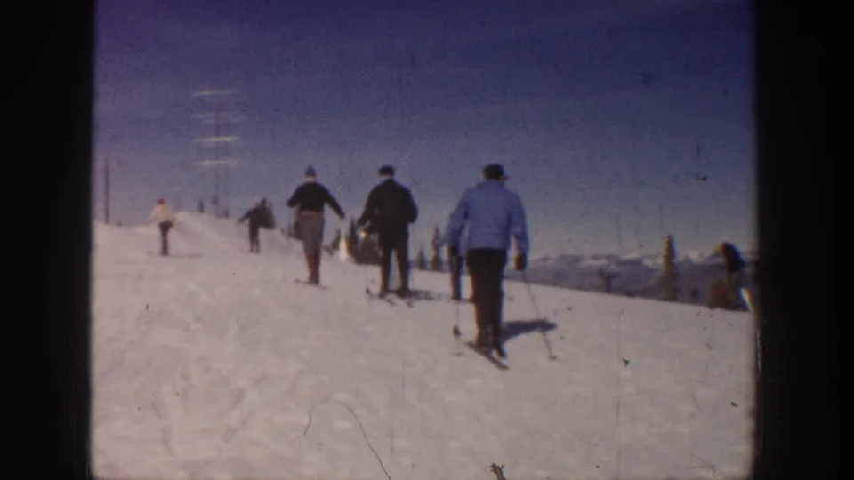 Skiers in 1961 Aspen home-movie footage walk up a groomed snowy slope, with distant mountains and lift poles or utility poles visible ahead.