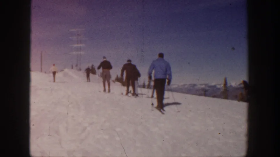 A group of skiers in period winter clothing walks across an Aspen snow slope, captured in warm, grainy 8mm color with distant ridges behind them.