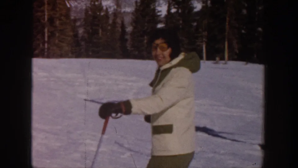 Adult woman in a white winter coat and sunglasses skiing on a snowy Aspen slope in 1961 home-movie footage.