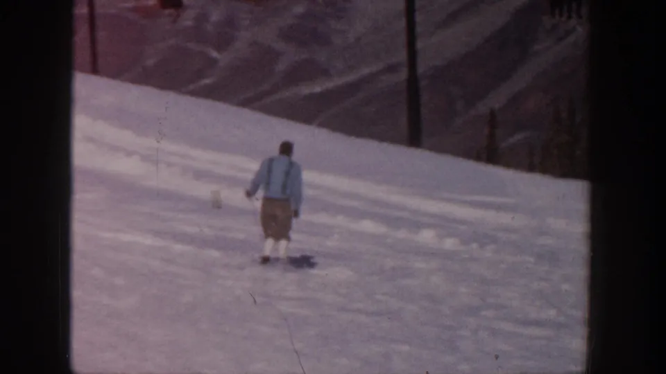 Adult skier in a blue jacket crosses the Aspen slope with mountain terrain and trees visible in the background.