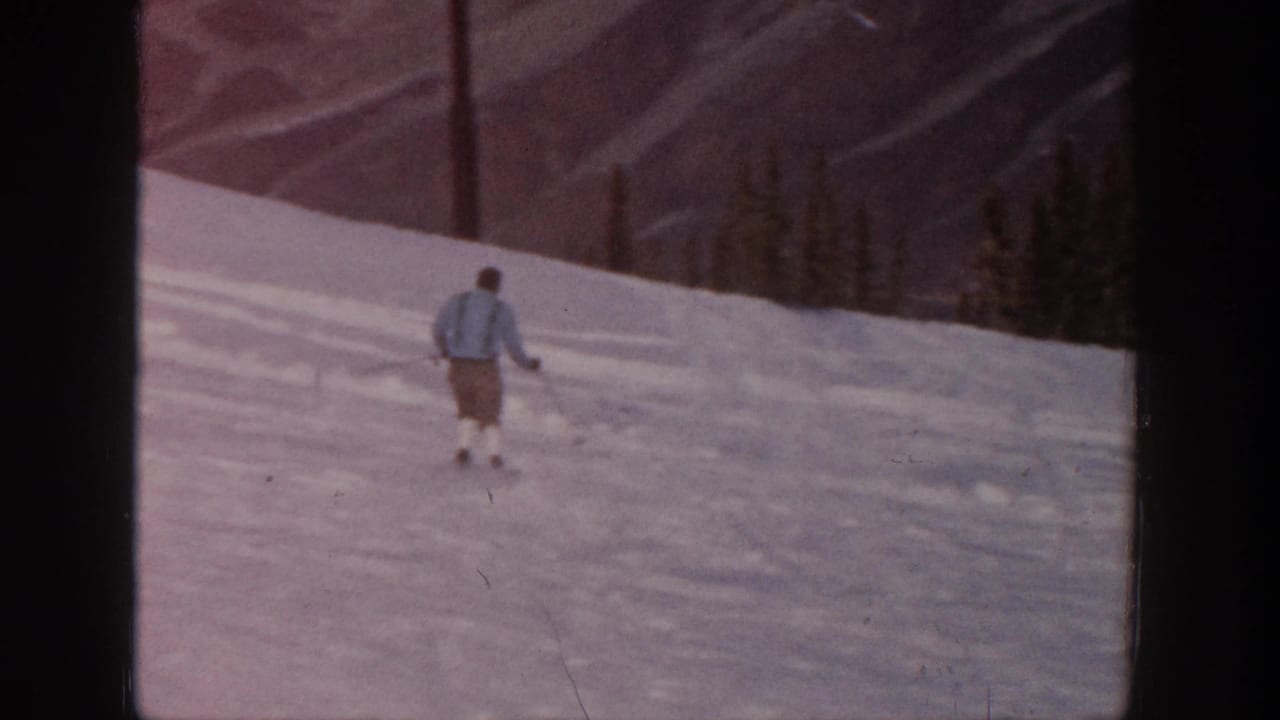 Adult skier in a light jacket moves down a gentle snowy slope in metadata-supported 1961 Aspen, with trees and mountains behind.