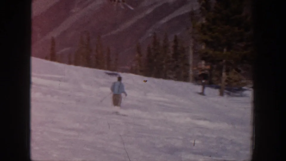 Distant adult skier traverses a snow-covered Aspen slope, with evergreens and muted mountain ridges visible in the background.