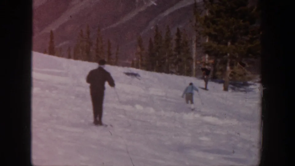 Two adult skiers appear on the snowy Aspen slope, one nearer in dark clothing and another farther ahead near the trees.