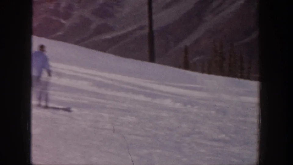 Adult skier in pale clothing glides on a snowy Aspen slope, framed by mountain ridges and the dark edge of the 8mm scan.