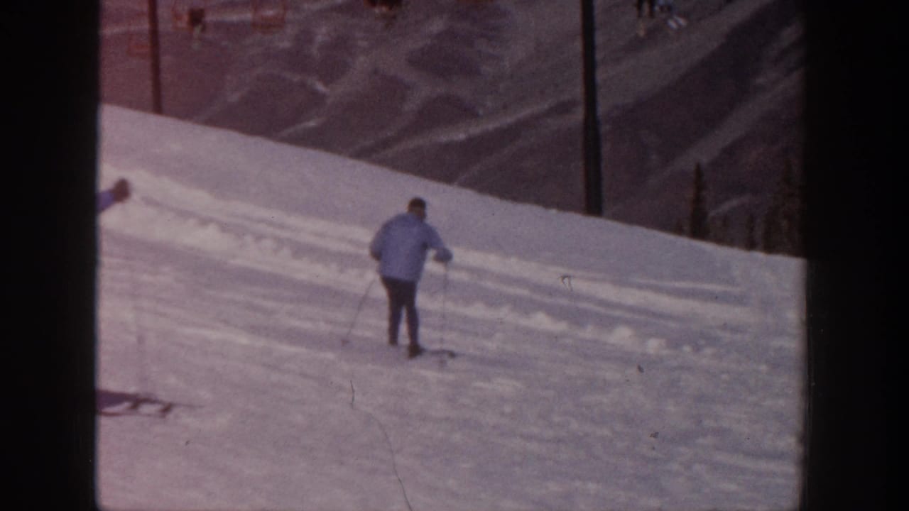 1961 Aspen home-movie frame showing two skiers on a snow slope, with lift poles, trees, and distant ridges behind them.