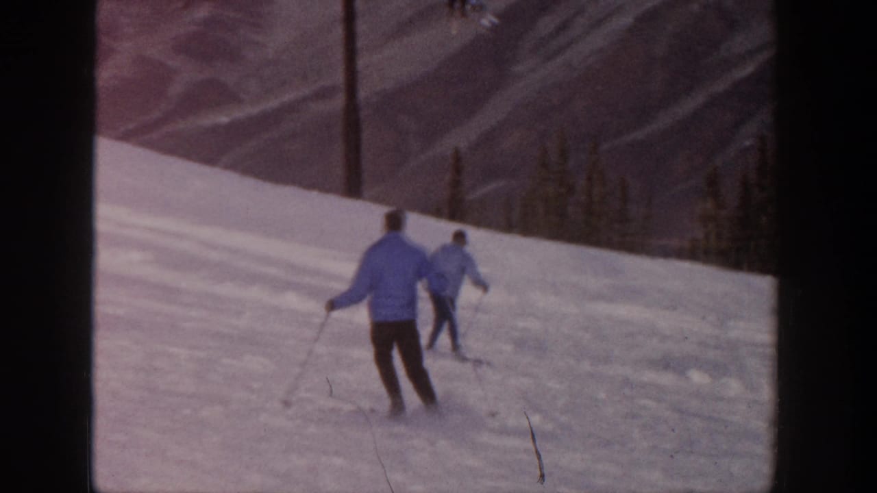 A foreground skier follows another skier across a sunlit snowy mountain slope.