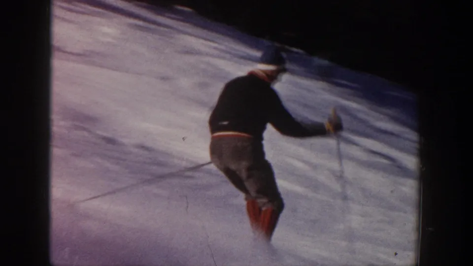 Rear view of the skier as he passes near the camera on an Aspen snow slope, with poles and red gaiter-like leg coverings visible.