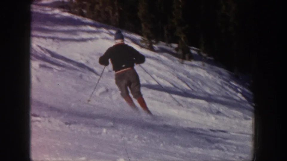 The skier continues downhill away from the camera on a snowy Aspen slope, with a dark treeline behind the run.