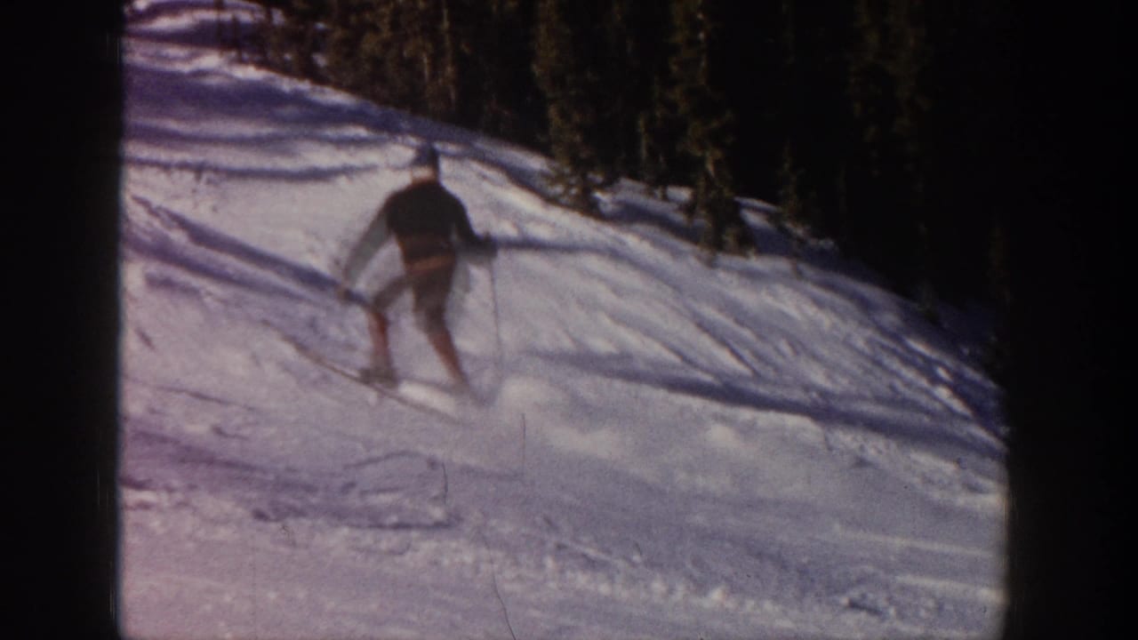 In Aspen, Colorado, 1961, a distant skier in dark upper clothing moves down a snow-covered slope bordered by evergreens.
