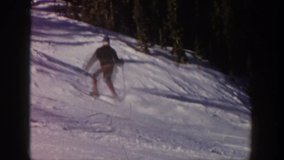 In Aspen, Colorado, 1961, a distant skier in dark upper clothing moves down a snow-covered slope bordered by evergreens.