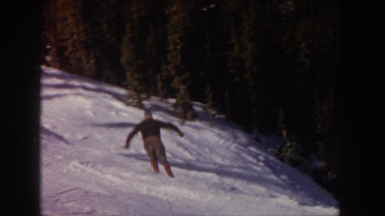 The skier continues across the Aspen slope, holding ski poles while turning below a dark line of evergreen trees.