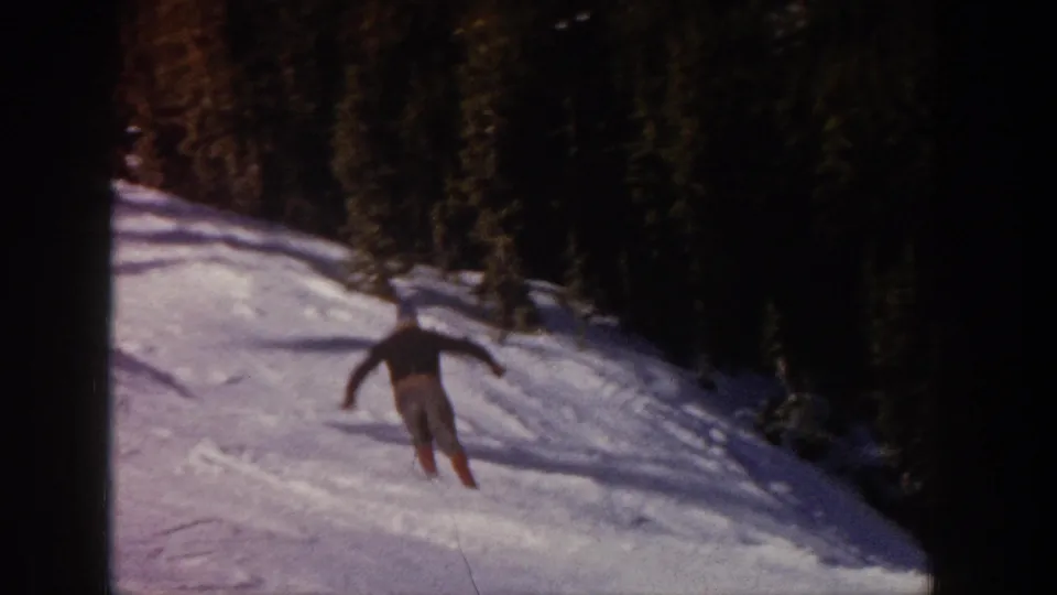 The skier continues across the Aspen slope, holding ski poles while turning below a dark line of evergreen trees.