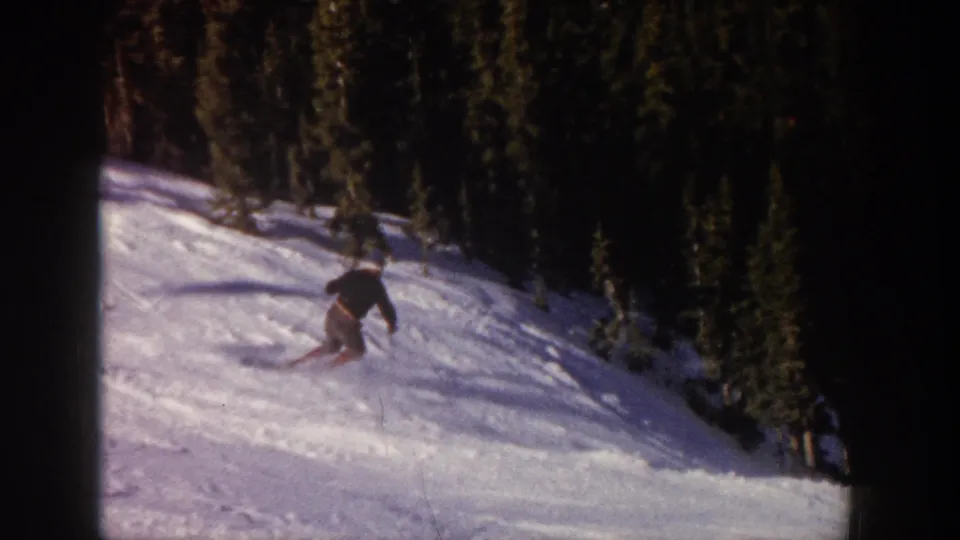 A skier in vintage 8mm Aspen footage bends into a turn on a bright snowy run with dense evergreens in the background.