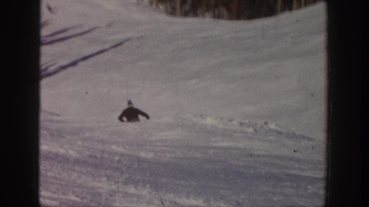 A small figure skis across an open snowy Aspen slope, framed by dark film borders and a line of winter trees.