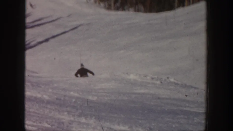 A small figure skis across an open snowy Aspen slope, framed by dark film borders and a line of winter trees.