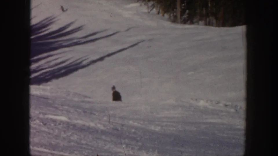 A distant skier continues downhill on a broad snowy Aspen slope, with long shadows and soft 8mm color visible across the frame.