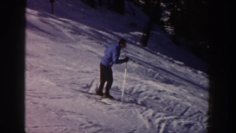 Adult skier in blue appears to slow or turn on the snowy Aspen slope, with trees and shadowed snow at the edge of frame.