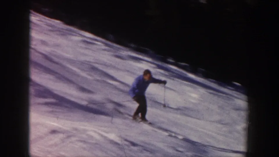 Adult skier crosses the Aspen snow slope in 1961 home-movie footage, framed against bright snow and a dark upper edge.