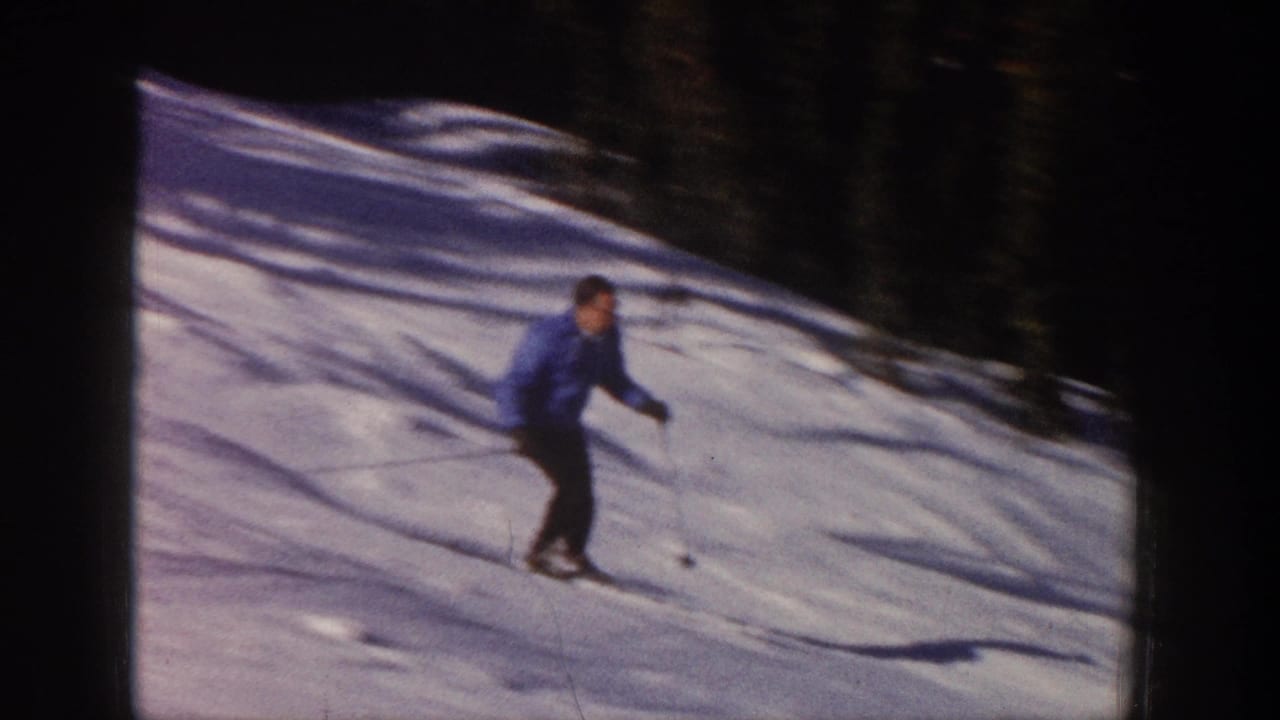 Skier in blue jacket glides across open snow with tree shadows behind him.