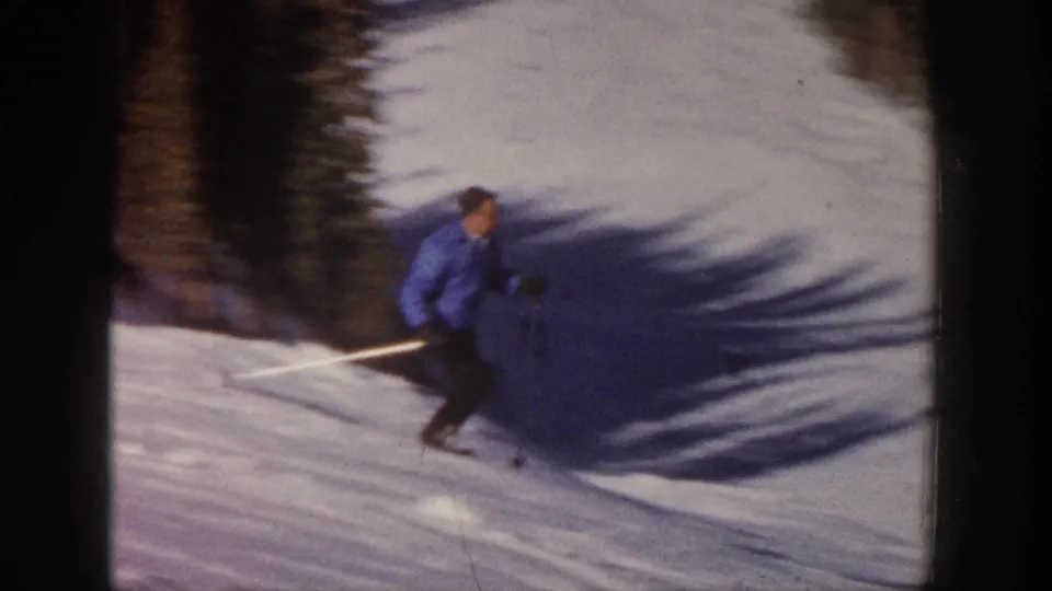 The skier leans into a turn on a snowy slope near the shadowed tree line in vintage Aspen 8mm footage.