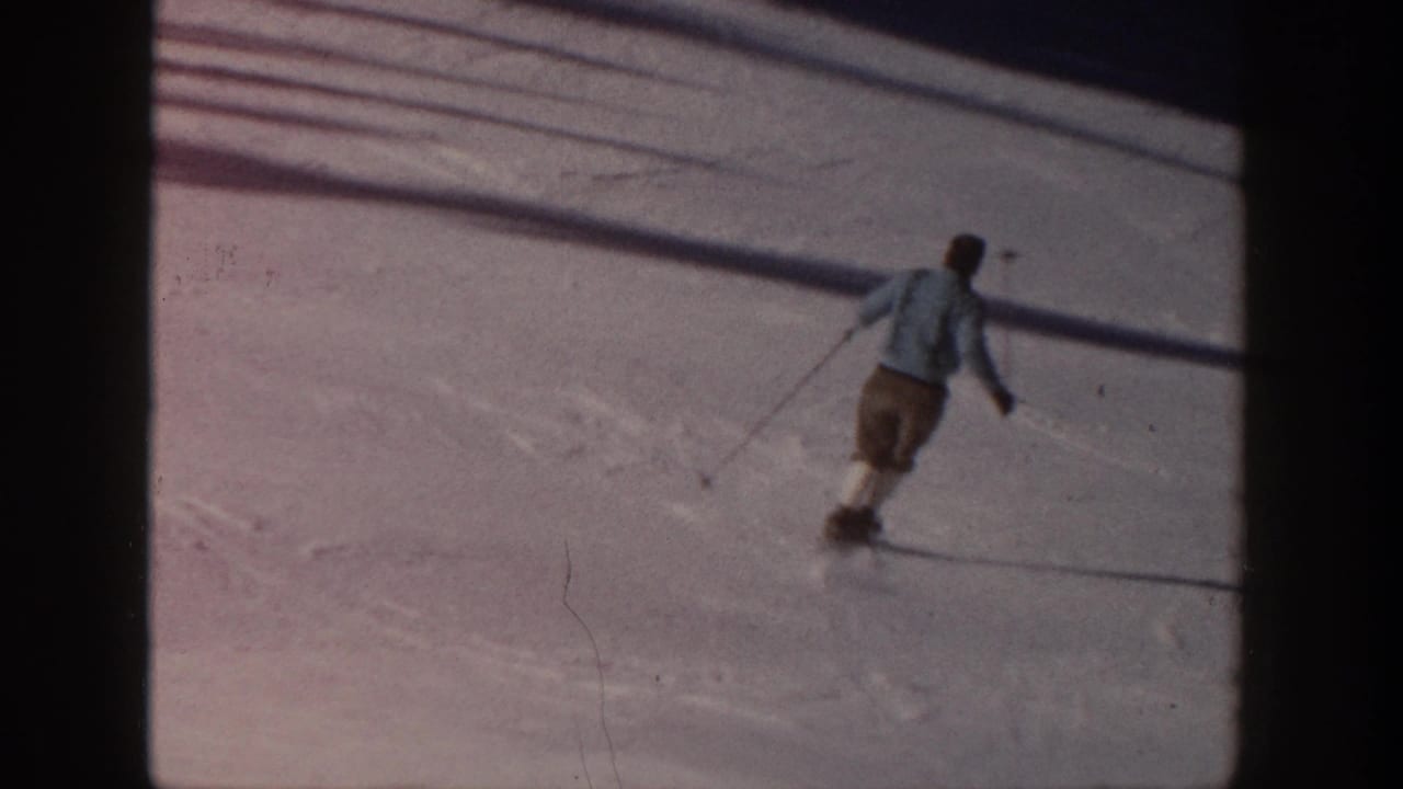 1961 Aspen home-movie frame showing a skier in a light jacket and brown shorts moving downhill across sunlit snow.