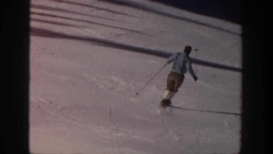 1961 Aspen home-movie frame showing a skier in a light jacket and brown shorts moving downhill across sunlit snow.