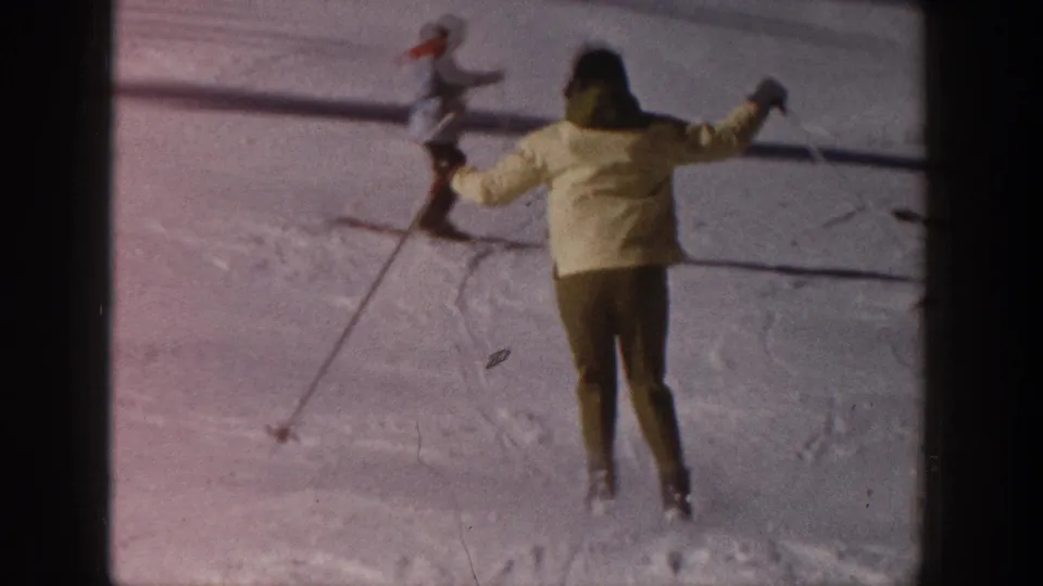 1961 Aspen frame showing a skier in a yellow jacket and dark pants skiing downhill with arms raised, with another skier nearby.