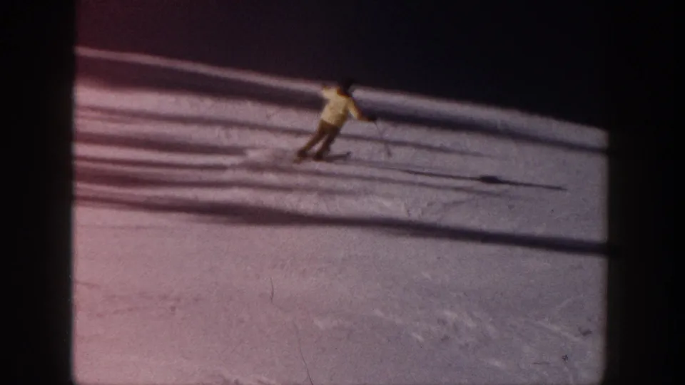 1961 Aspen footage frame showing the skier angling downhill through tracked snow and strong slope shadows.