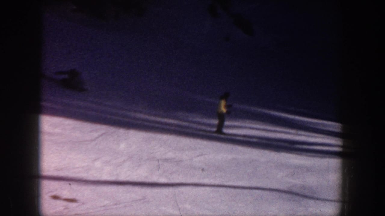 Skier in a yellow coat stands or glides on a snowy slope with long shadows.