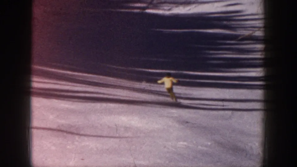 1961 Aspen home-movie frame with a yellow-coated skier moving downhill across snow marked by shadows and film scratches.