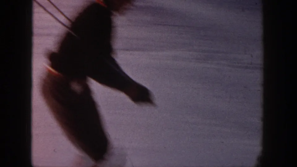 A skier passes near the 8mm camera in soft motion blur, with snow and shadowed terrain behind.