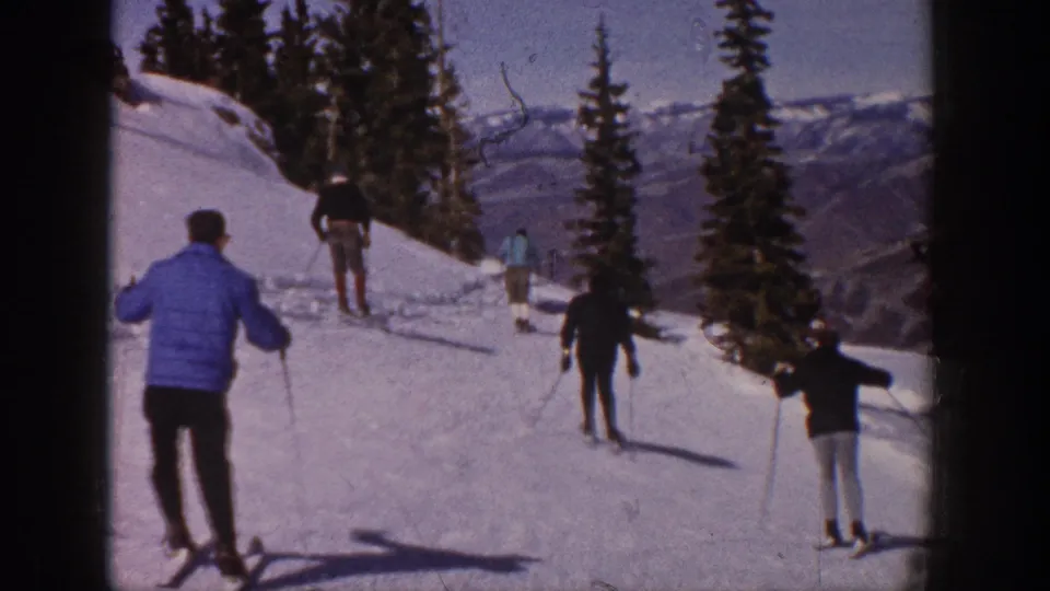 A blue-jacketed skier follows a small group downhill on a snowy Aspen trail lined with evergreen trees.