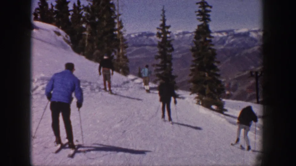 Skiers descend an Aspen slope in 1961 home-movie footage, with trees, distant ridges, and a dark edge of a structure at right.