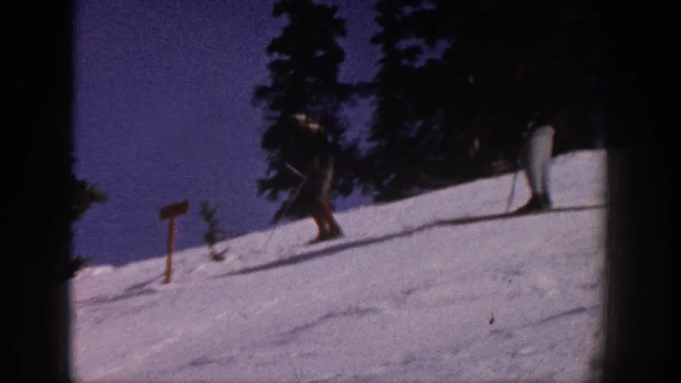 1961 Aspen frame with a skier on a snowy hillside, evergreen trees, and a small trail sign under blue sky.