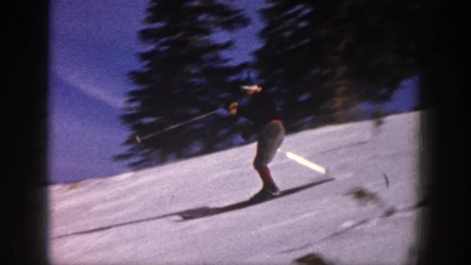 1961 Aspen frame showing a skier turning downhill on bright snow with evergreen trees behind.