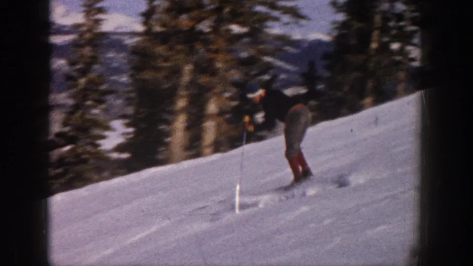 The skier continues downhill on a bright snowy Aspen slope, with evergreen trees and distant mountains visible in the 8mm frame.
