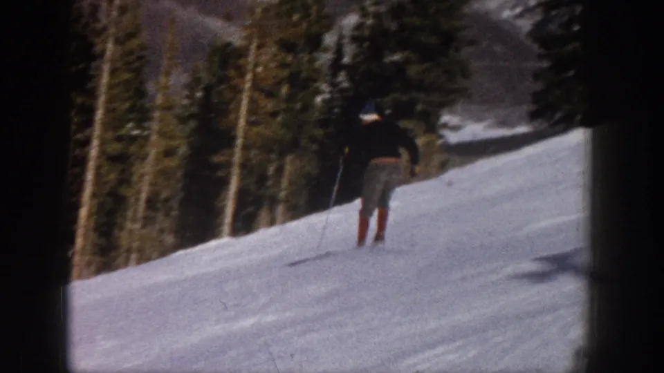 A rear view of the skier moving away across a snowy 1961 Aspen mountain slope, with trees lining the background.