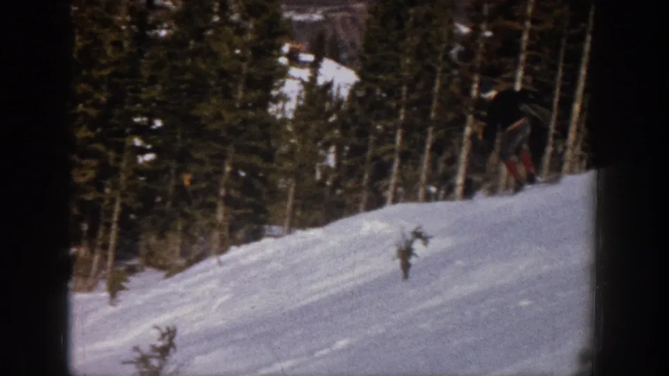 The skier appears near the right edge of the frame on a snowy Aspen slope, with trees and small foreground shrubs visible.