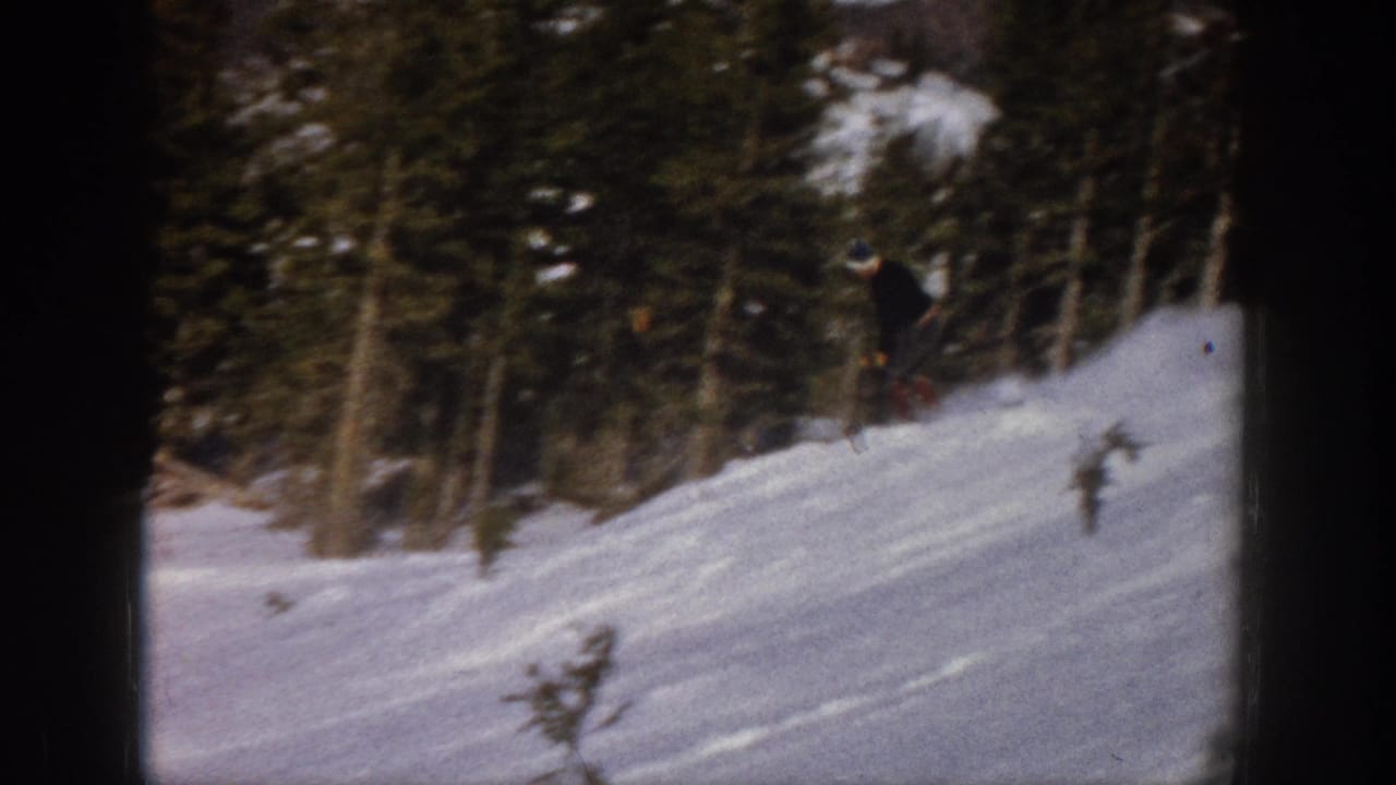 In supplied 1961 Aspen 8mm footage, a distant skier in dark clothing crosses a bright snowy slope near evergreen trees.