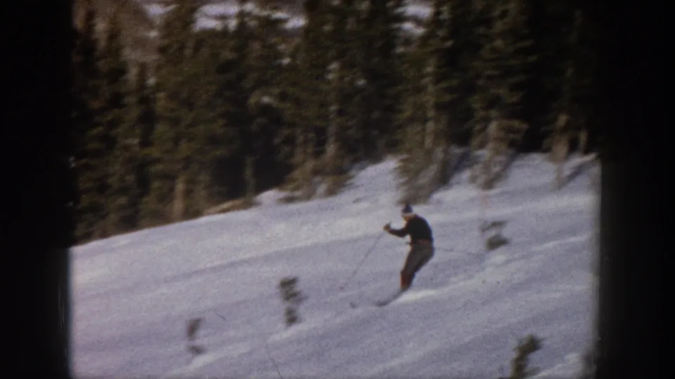 A skier descends an open Aspen snow slope with poles in hand, framed by dark evergreens in grainy 8mm color.