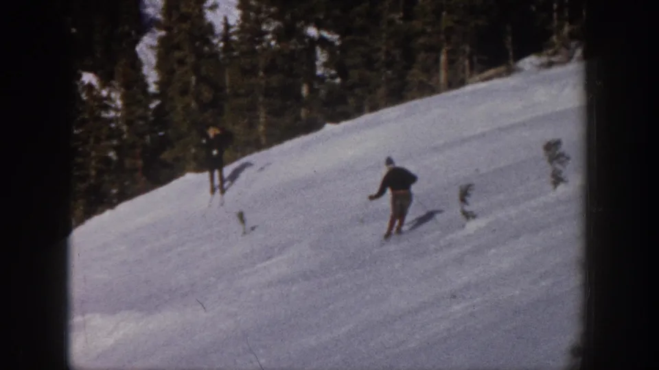 In the Aspen home-movie scene, the skier is seen from behind moving downhill while another figure stands near the treeline.