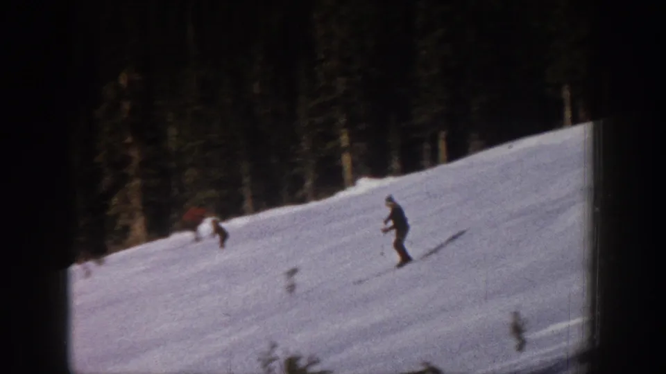 1961 Aspen 8mm frame with two distant skiers crossing a snowy mountain slope below evergreen trees.