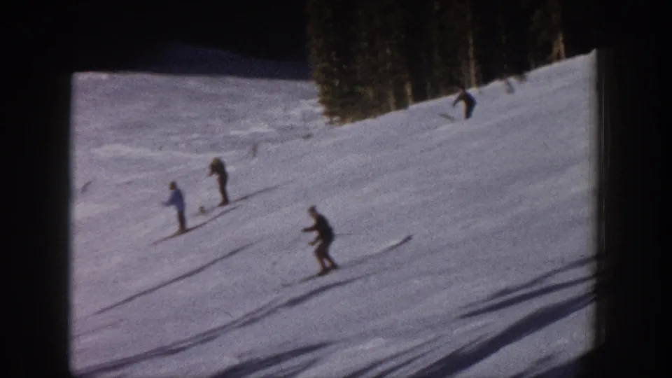 1961 Aspen home-movie frame showing several skiers spread across a snowy slope under an evergreen treeline.
