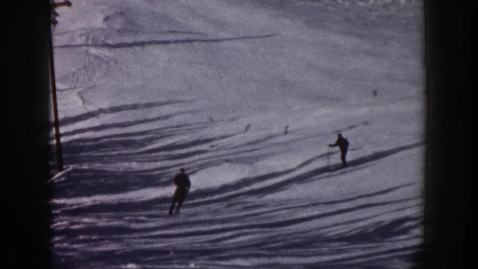 Distant adult skiers descend a snowy Aspen slope in 1961, framed by long shadows and the soft grain of color 8mm home-movie film.