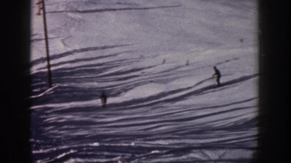 A distant skier moves downhill on a snowy Aspen slope, with elongated shadows and 8mm film edges shaping the archival home-movie frame.